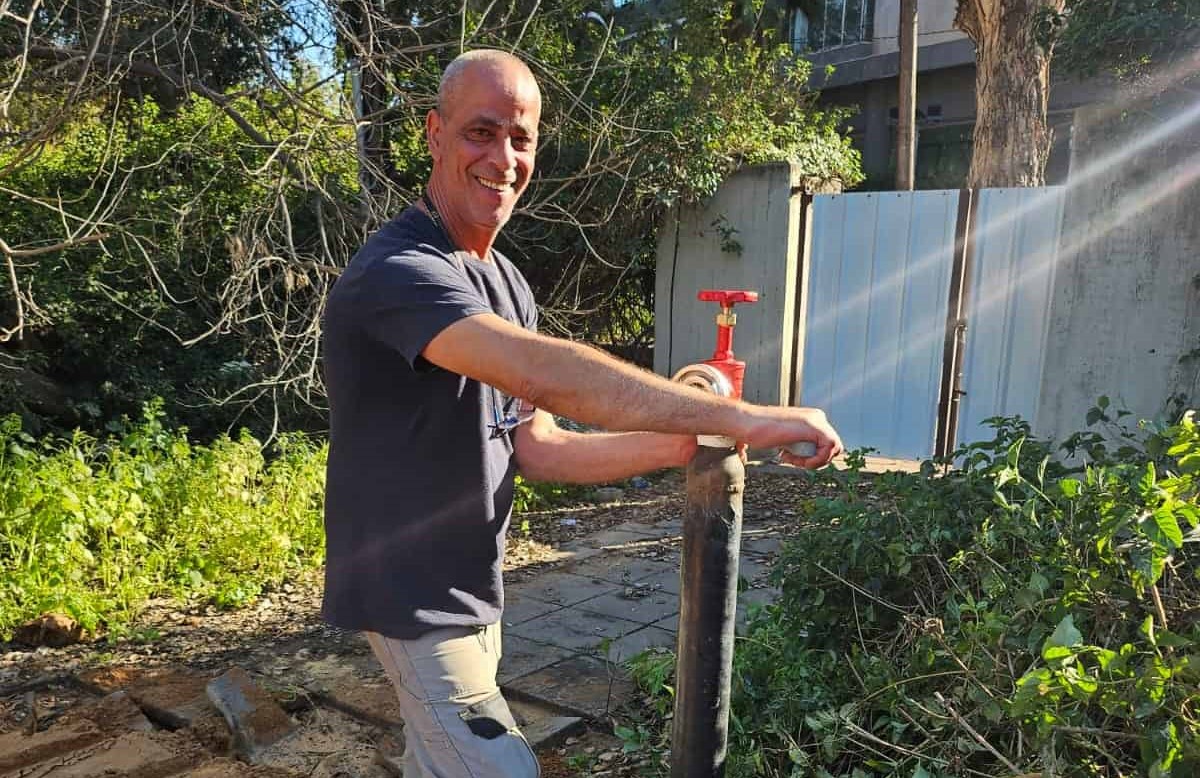 גבר מחייך עומד בחוץ, מסובב ברז בעל ידית אדומה המחובר לצינור שחור אנכי. הוא מוקף בצמחייה ירוקה, ואור השמש מסנן מבעד לעצים שמאחוריו. | עמית נעם תמיד בזמן - אינסטלוטור בתל אביב
