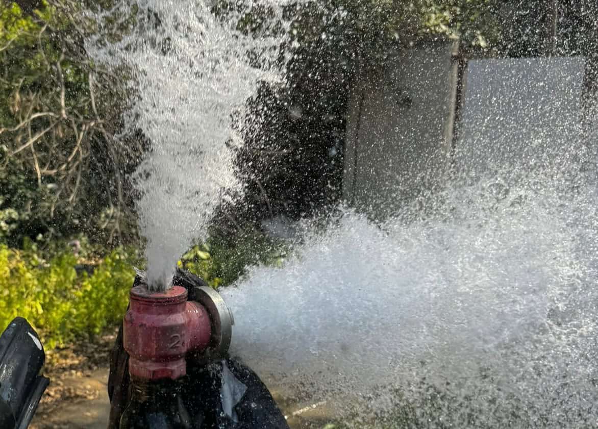 תקריב של מים המתיזים בעוצמה מתוך צינור או ברז אדום בחוץ, ויוצרים סילון של טיפות לבנות, כשברקע המטושטש נראים צמחייה וקיר לבן. | עמית נעם תמיד בזמן - אינסטלוטור בתל אביב