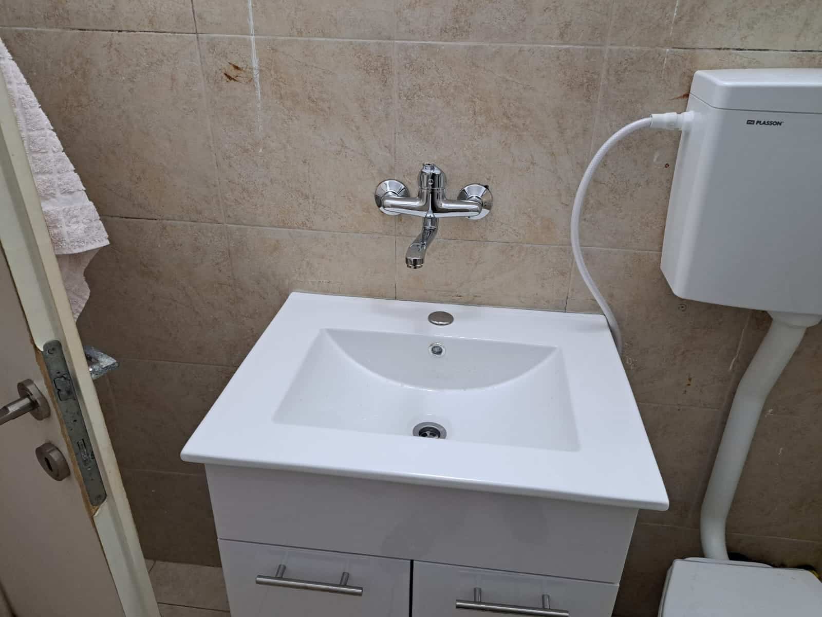 כיור לבן קטן עם ברז כסוף מותקן על קירות מחופים באריחים בצבע בז'. משמאל תלוי מגבת, ומימין נמצא מיכל הדחה לבן לאסלה. מתחת לכיור יש שתי מגירות. | עמית נעם תמיד בזמן - אינסטלוטור בתל אביב