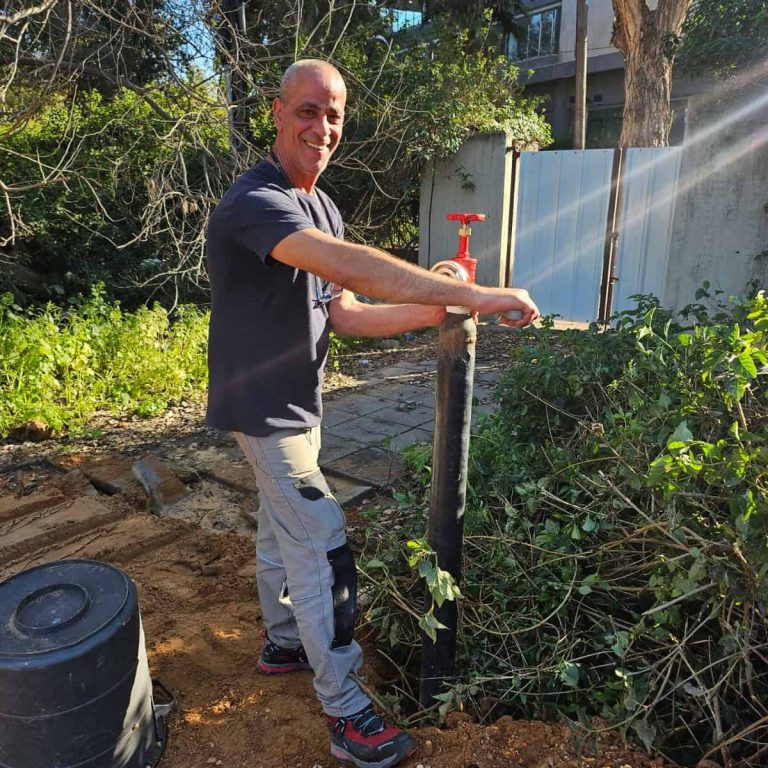 גבר מחייך בזמן שהוא מסובב ברז אדום על צינור שחור בחוץ, מוקף בצמחייה ובאדמה, כשברקע נראים מבנים ועצים. | עמית נעם תמיד בזמן - אינסטלוטור בתל אביב