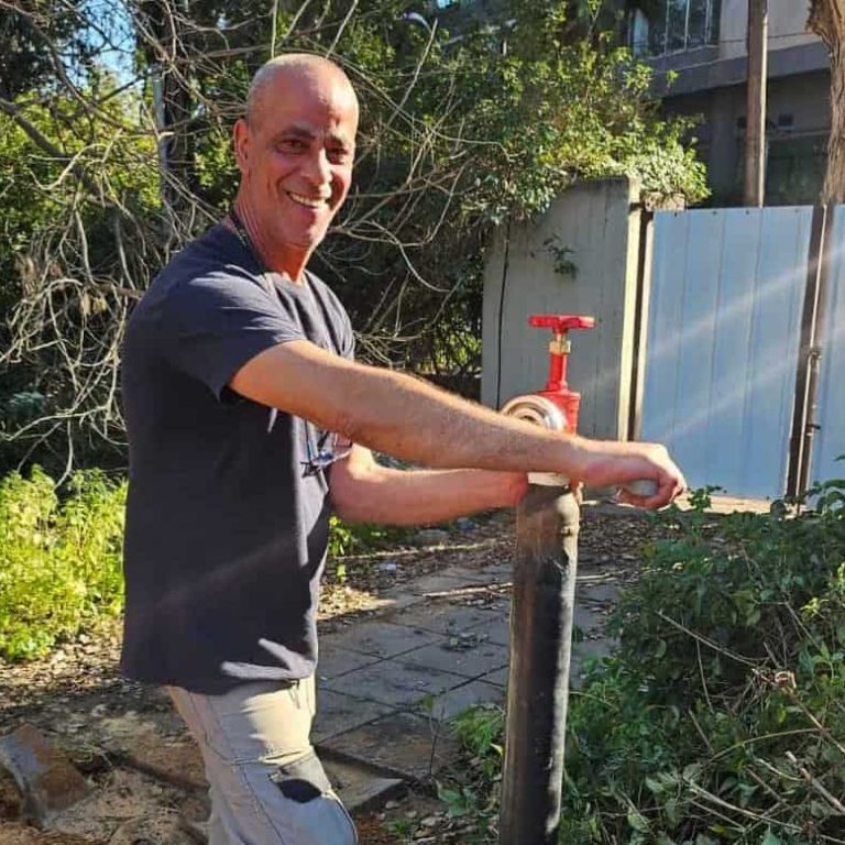 גבר מחייך עומד בחוץ, מסובב ברז בעל ידית אדומה המחובר לצינור שחור אנכי. הוא מוקף בצמחייה ירוקה, ואור השמש מסנן מבעד לעצים שמאחוריו. | עמית נעם תמיד בזמן - אינסטלוטור בתל אביב